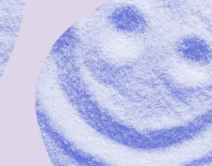 Grafika, w tle piasek na którym jest narysowany uśmiech z fioletowymi elementami graficznymi. Medicante